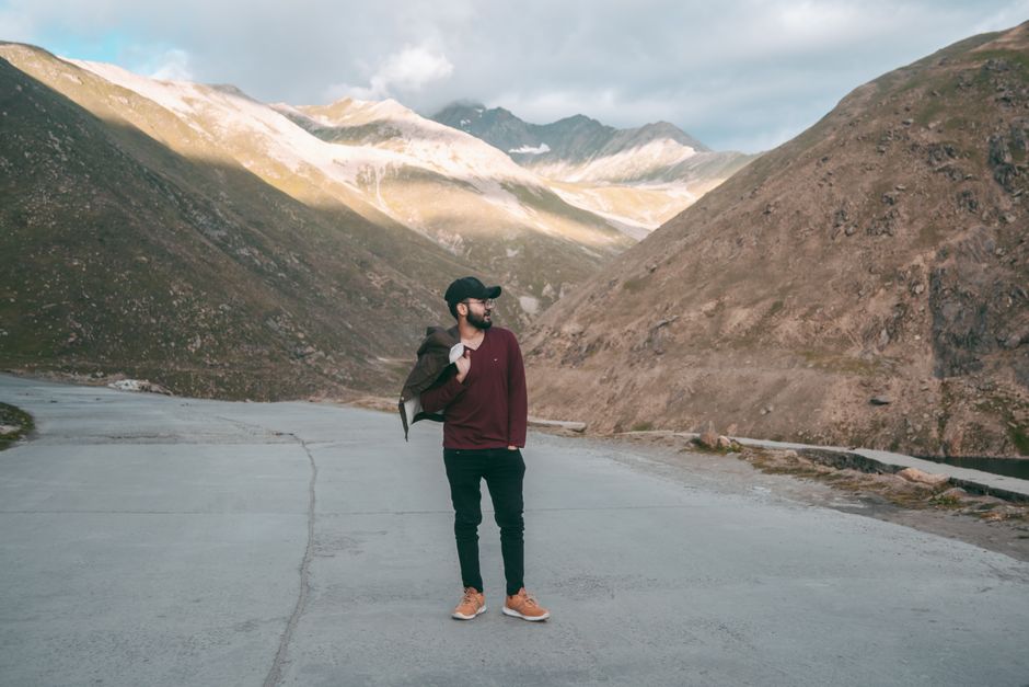 A man stands on a concrete road amidst a stunning mountainous landscape under a cloudy sky