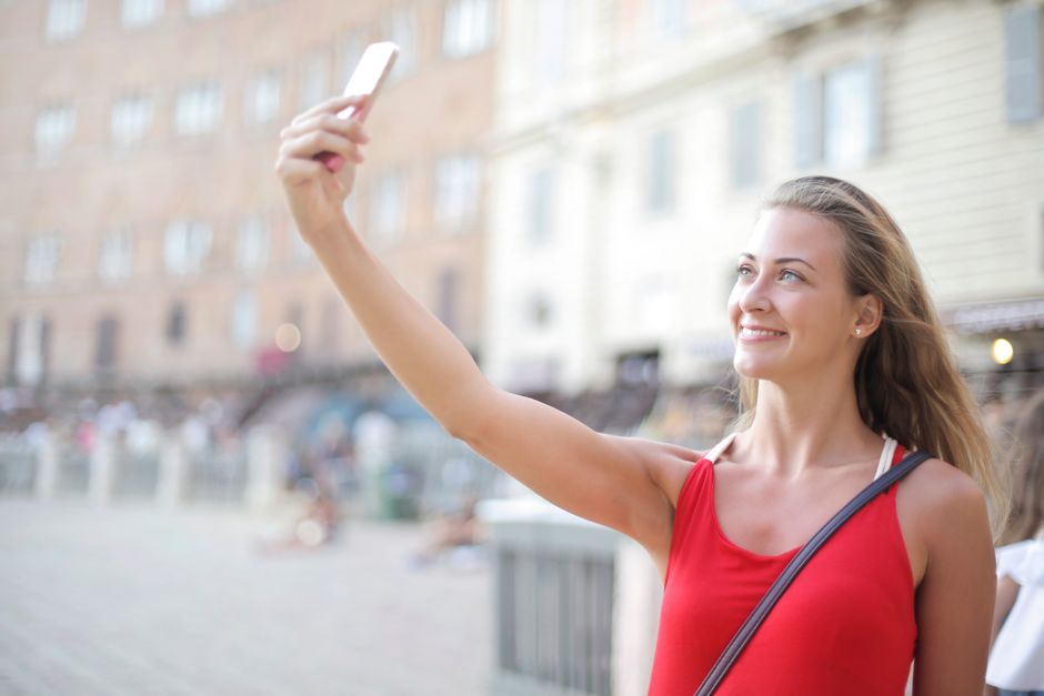 Woman in red top smiling while taking a selfie in an urban setting.