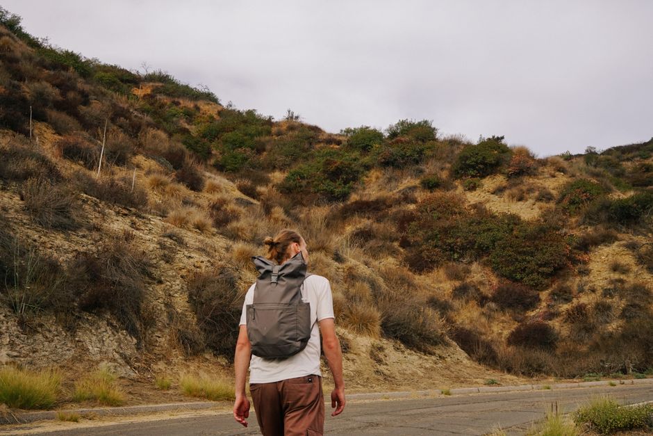 A man with a backpack walks uphill on a cloudy day, exploring a dry and hilly landscape.