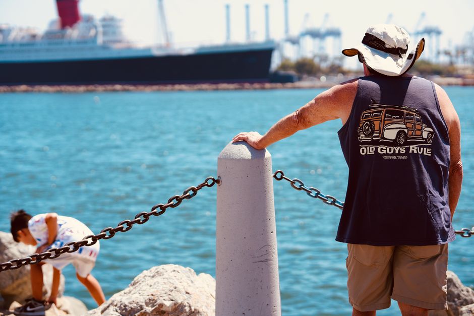 Man admires ocean view near port, child plays on rocks, ship in background.