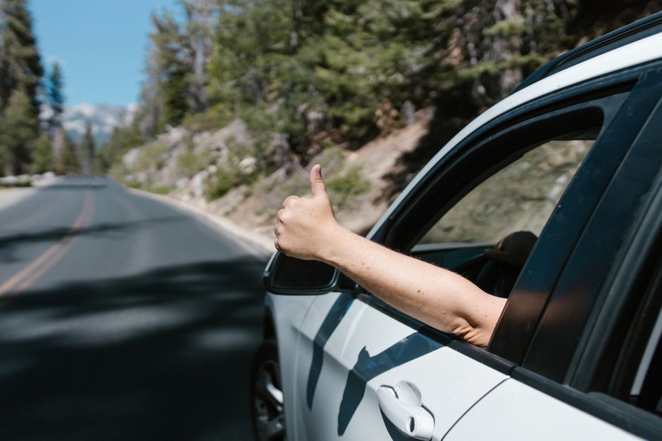 A person gives a thumbs up gesture from a car window while driving on a scenic forest road.