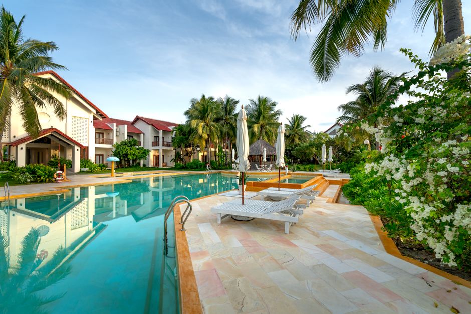 A serene poolside view with sunloungers and villas surrounded by palm trees and lush greenery.