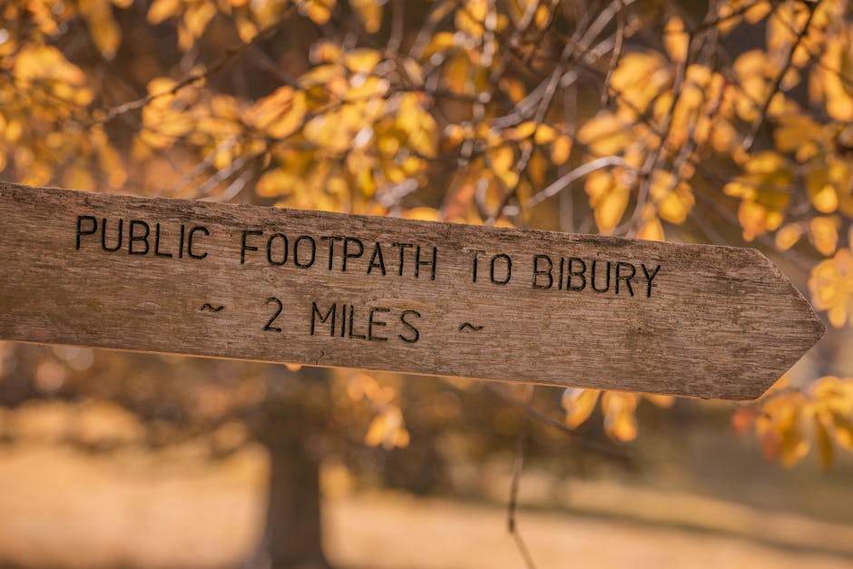 Rustic wooden signpost pointing to Bibury in the Cotswolds, England, surrounded by autumn leaves.