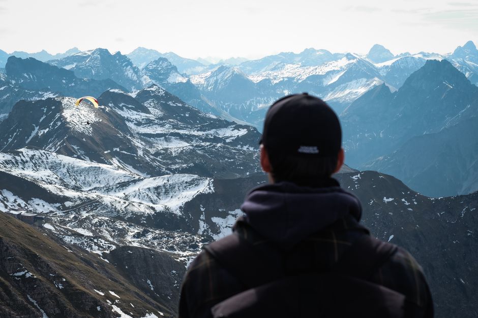 A person in a cap views snowy mountain peaks and a paraglider in the distance.