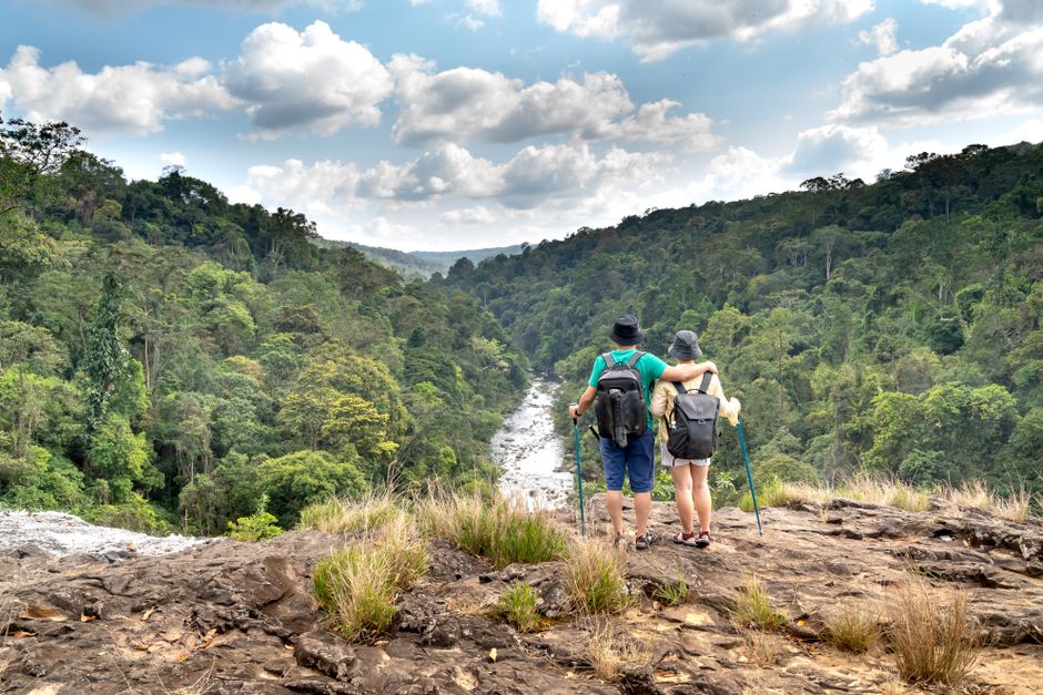 A couple enjoys a summer hike, overlooking lush green mountains and a flowing river.