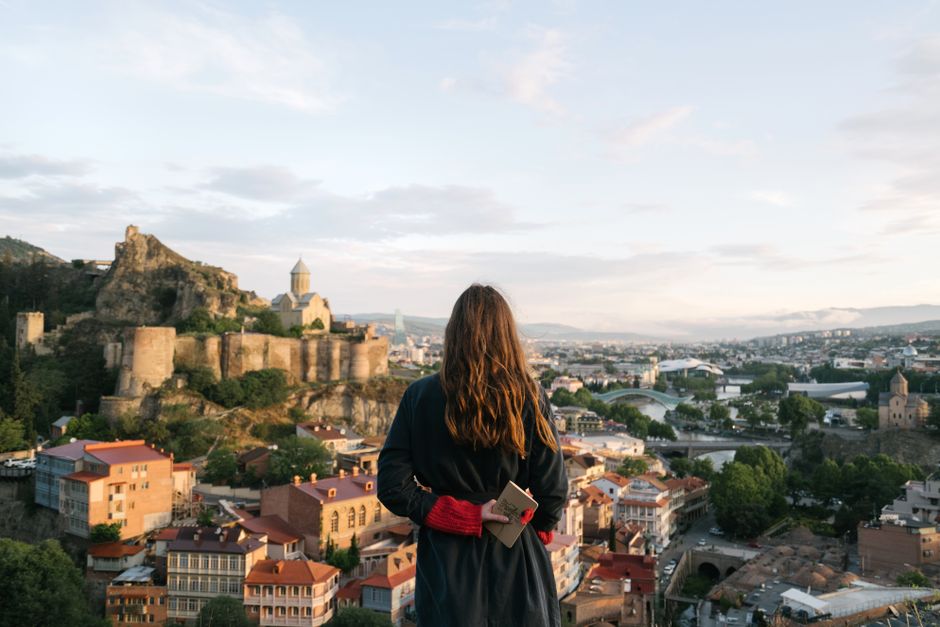 A woman stands with a notebook, gazing at the Tbilisi skyline and historic fortress, capturing a moment of nostalgia.