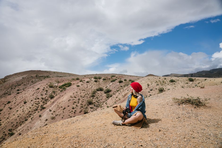 Woman in red beanie sitting in a desert landscape with rocky mountains and blue sky.