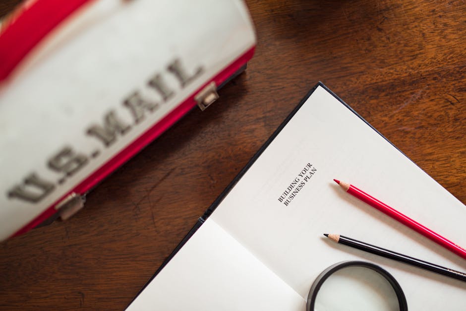 Open book with 'Building Your Business Plan' text, pencils, and U.S. Mail bag on a wooden desk.