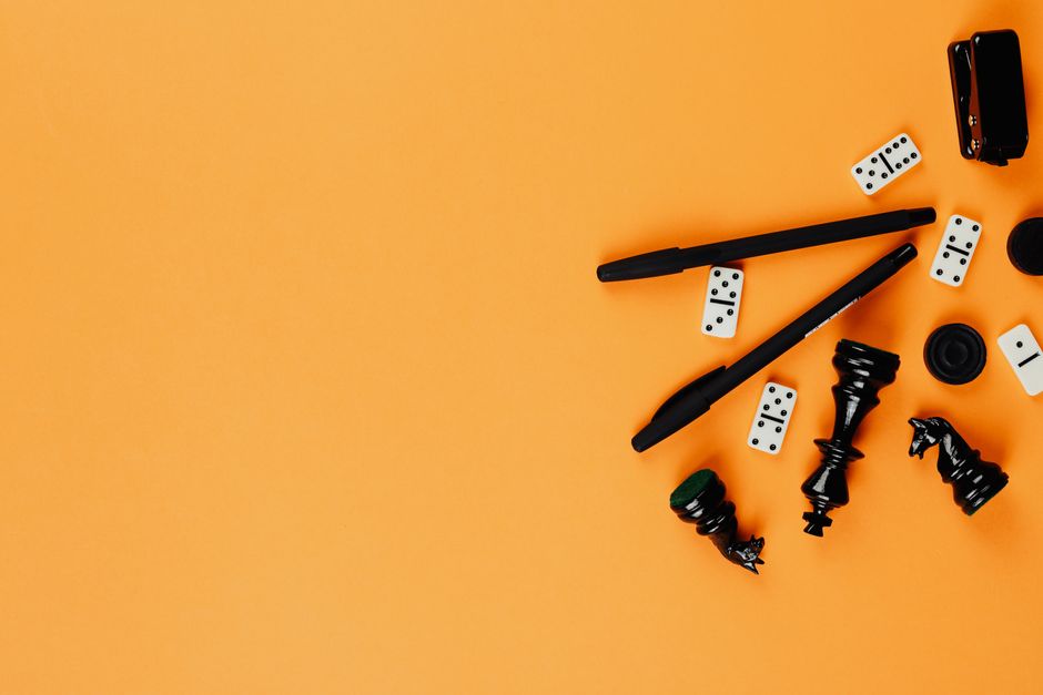 A minimalist arrangement of chess pieces, dominoes, pens, and a stapler on a vibrant orange background.