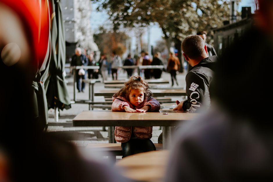 A father and daughter sitting at an outdoor cafe as people walk by on a sunny day.