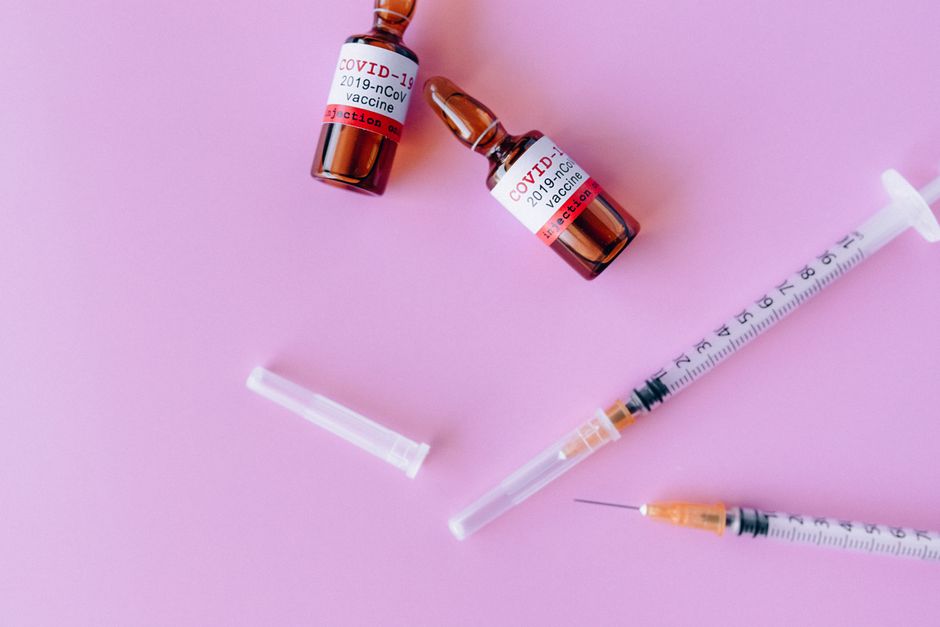 Flat lay of COVID-19 vaccine vials and syringe on a pink surface, representing global health efforts.