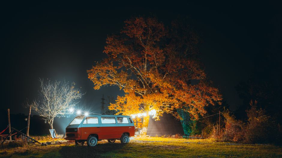A vintage red camper van parked under a brightly lit autumn tree on a serene night.