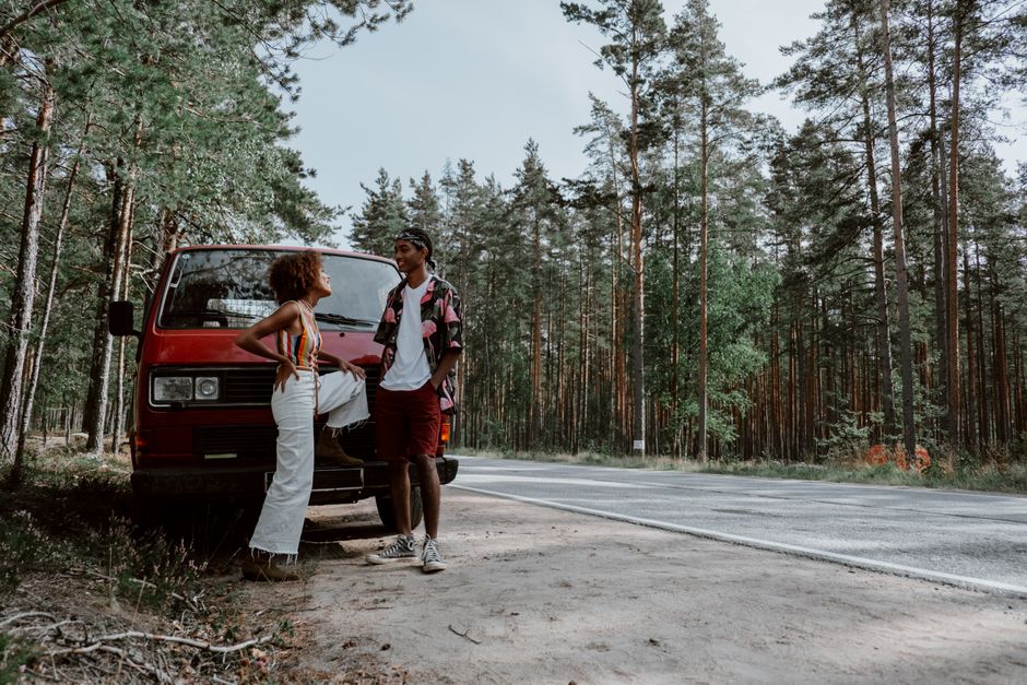 Two people relaxing by a vintage red van in a pine forest, embodying adventure and travel spirit.