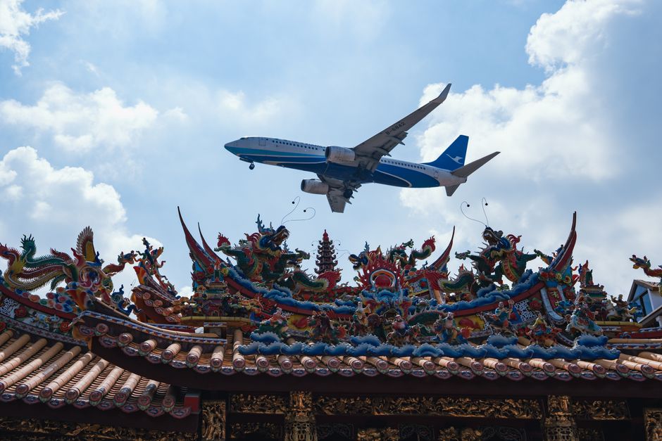 Airplane flying over a traditional Chinese temple rooftop in Xiamen, China under a bright blue sky.